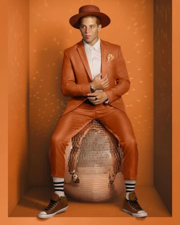 Man in orange suit sitting on disco ball in orange photo studio. He wears a matching hat and brown sneakers with striped socks.